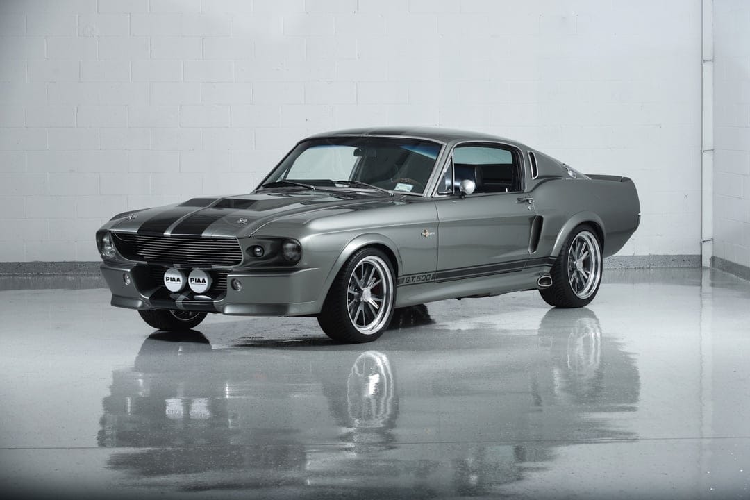 Silver Shelby GT500 Eleanor Mustang with black stripes parked on a reflective studio floor by a white brick wall.
