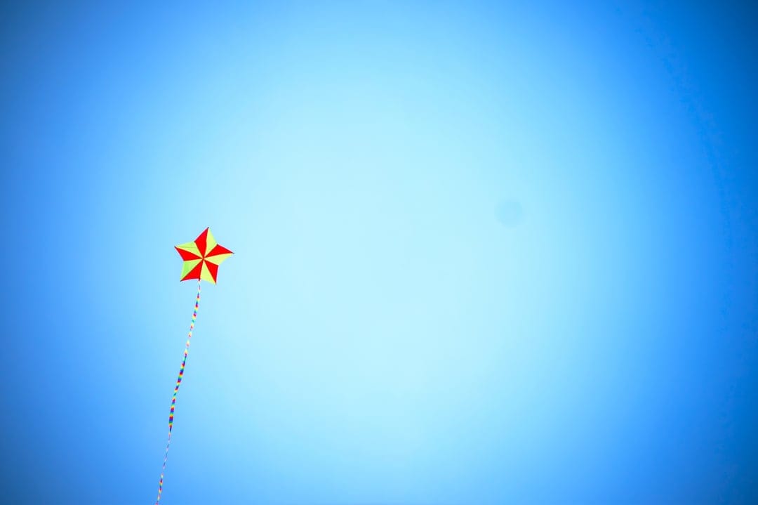 Star-shaped kite with red and yellow panels flying high in a clear blue gradient sky with a long trailing tail.