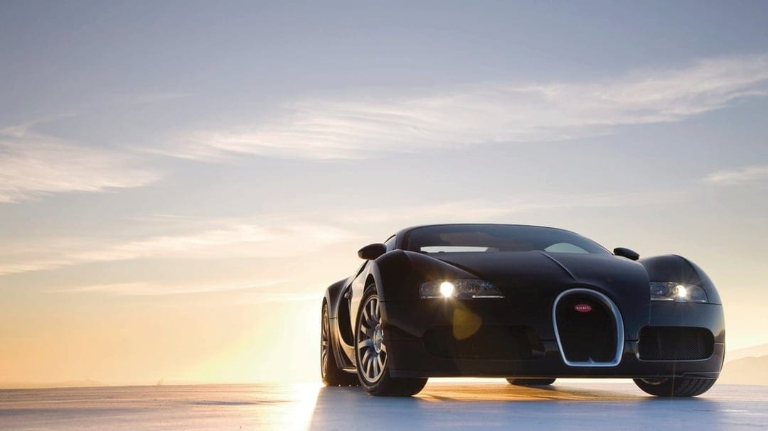 Front view of a black Bugatti Veyron supercar with headlights on against a soft blue and gold sunset sky.