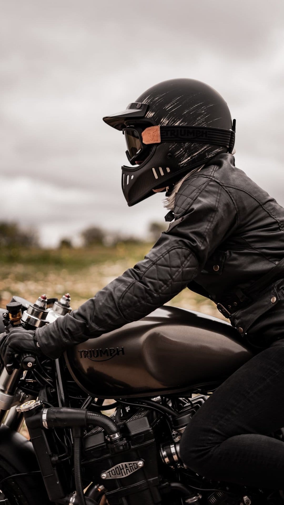Rider in a black quilted jacket and distressed helmet on a dark Triumph motorcycle against a blurred field.