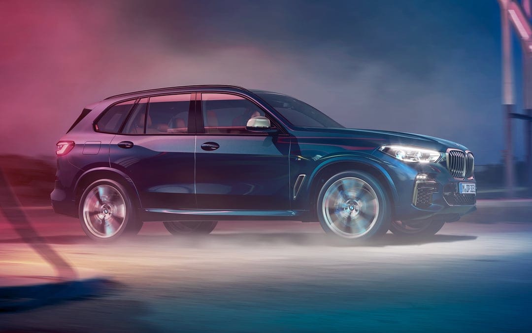 Blue BMW X5 SUV driving fast with motion blurred wheels through pink and blue mist on a dark nocturnal road.