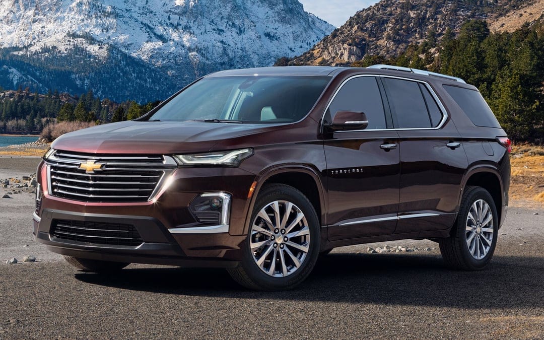 Dark brown Chevrolet Traverse SUV parked on a road with snowy mountain peaks and pine trees in the background.
