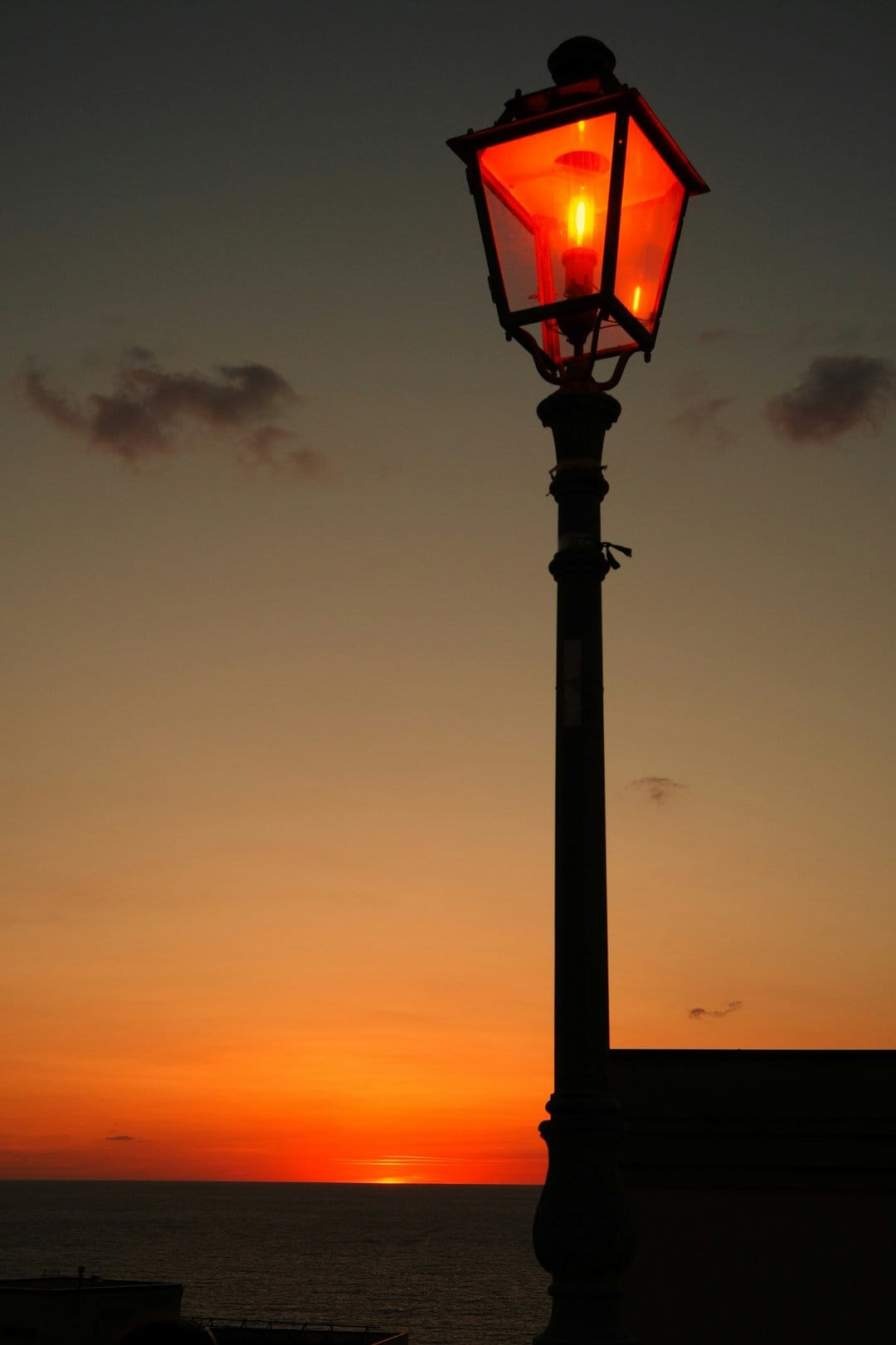 Antique metal lantern glowing with warm orange light against a dramatic sunset sky over the calm ocean horizon.