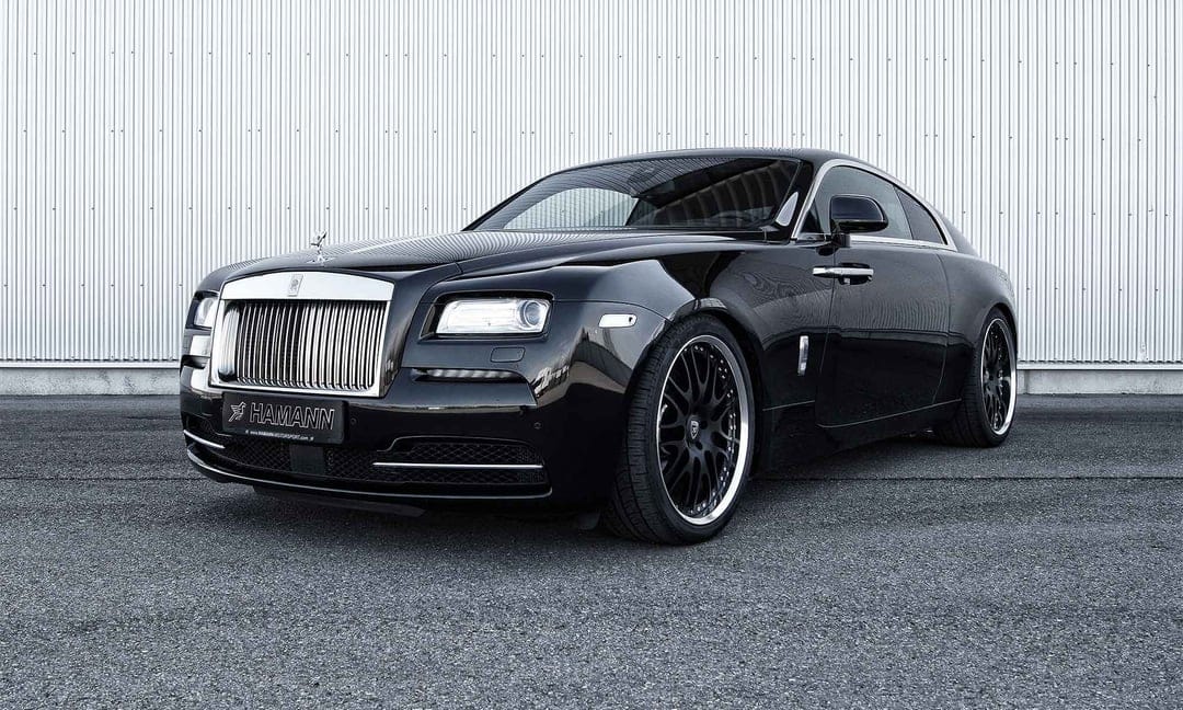 Black Rolls-Royce Wraith with Hamann wheels parked against a ribbed metallic wall under diffused lighting.