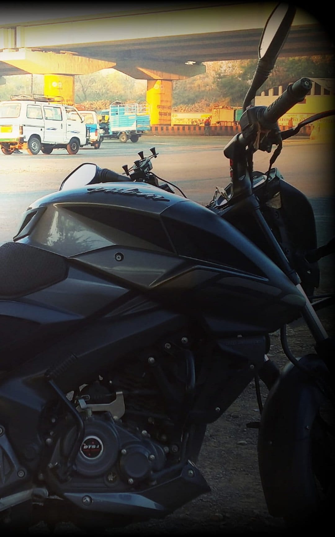 Dark motorcycle engine and fuel tank reflecting sunlight with blurred trucks under a concrete city overpass.