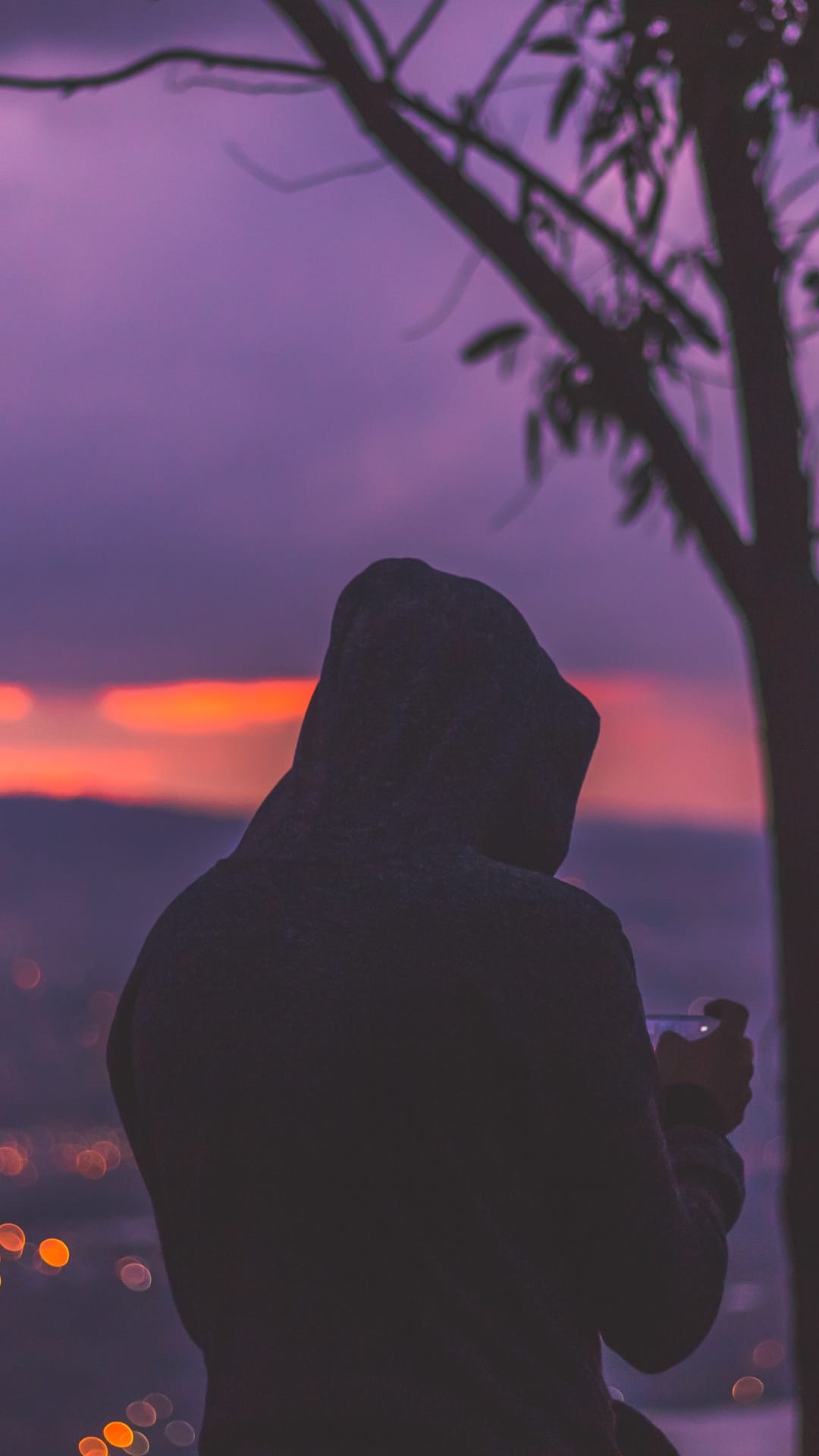 Hooded person stands against a purple sunset sky overlooking blurred city lights from an urban overlook.