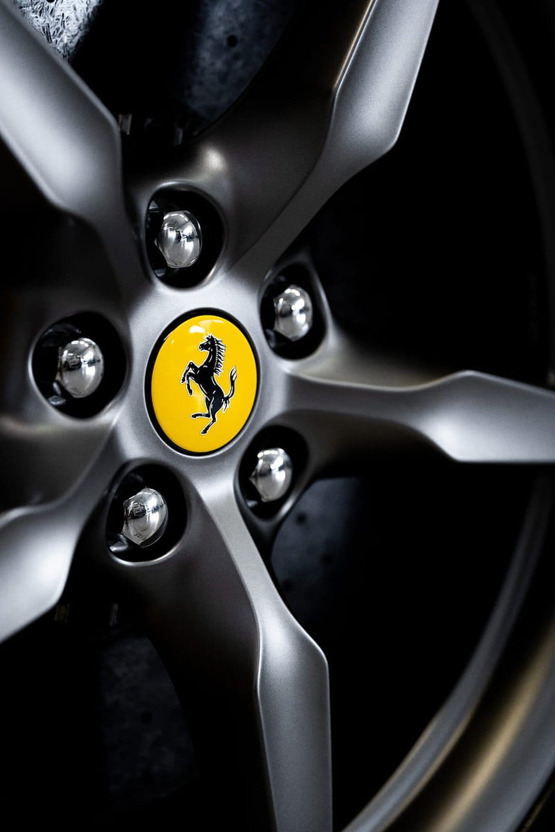 Ferrari prancing horse emblem at the center of a silver alloy wheel with polished lug nuts on a dark background.