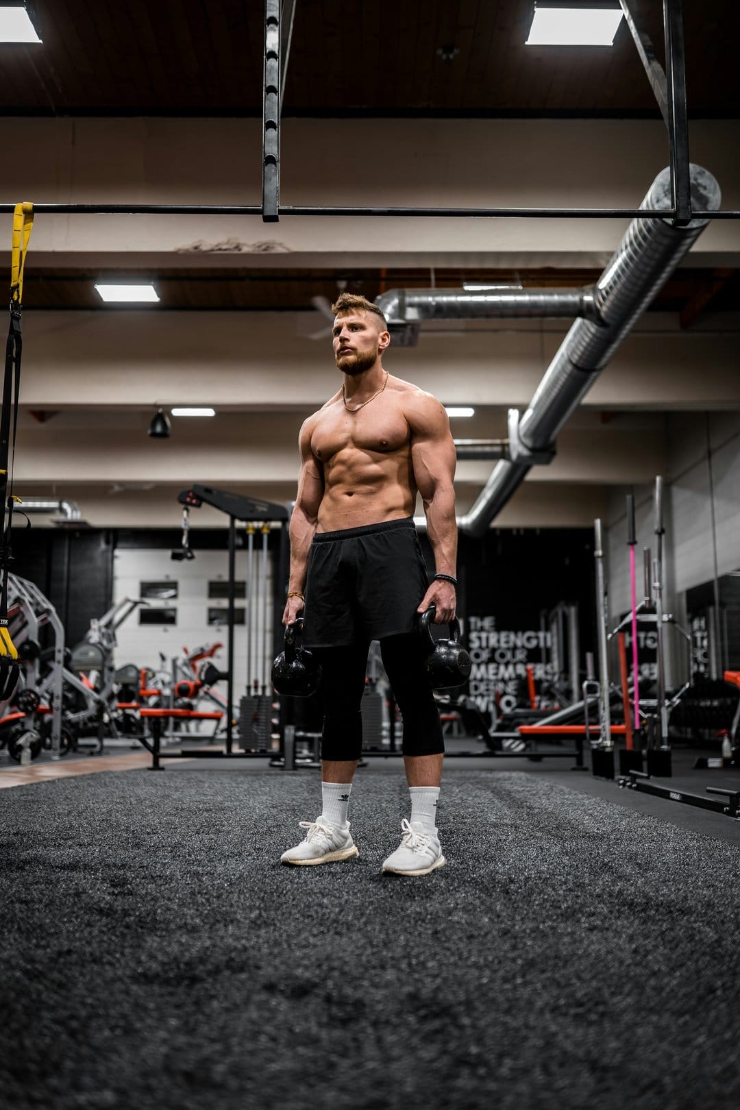 Muscular man with defined abs holding two kettlebells in a gym while wearing black shorts and white sneakers.