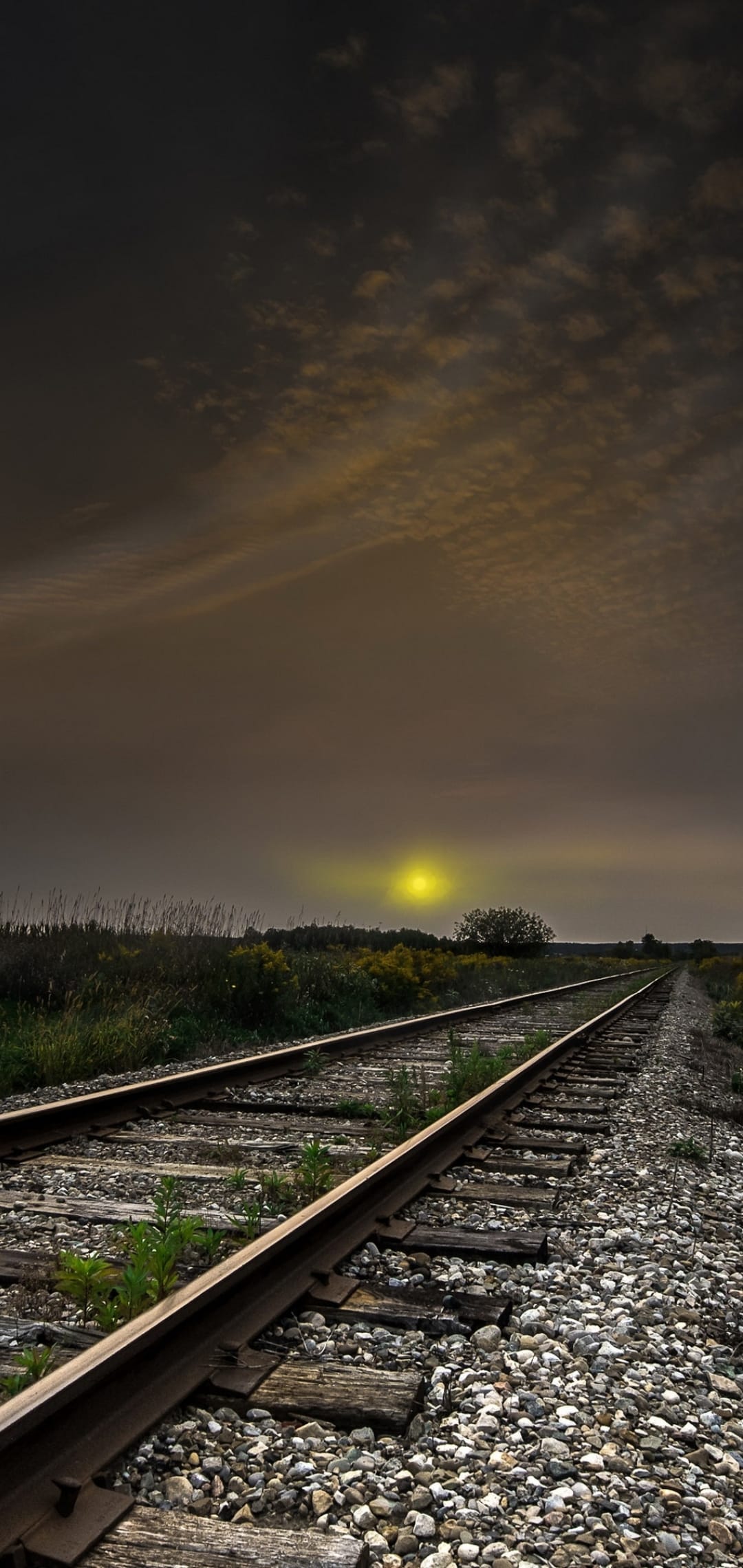 Weathered railroad tracks stretch into a hazy distance beneath a thick layer of glowing golden clouds at dusk.