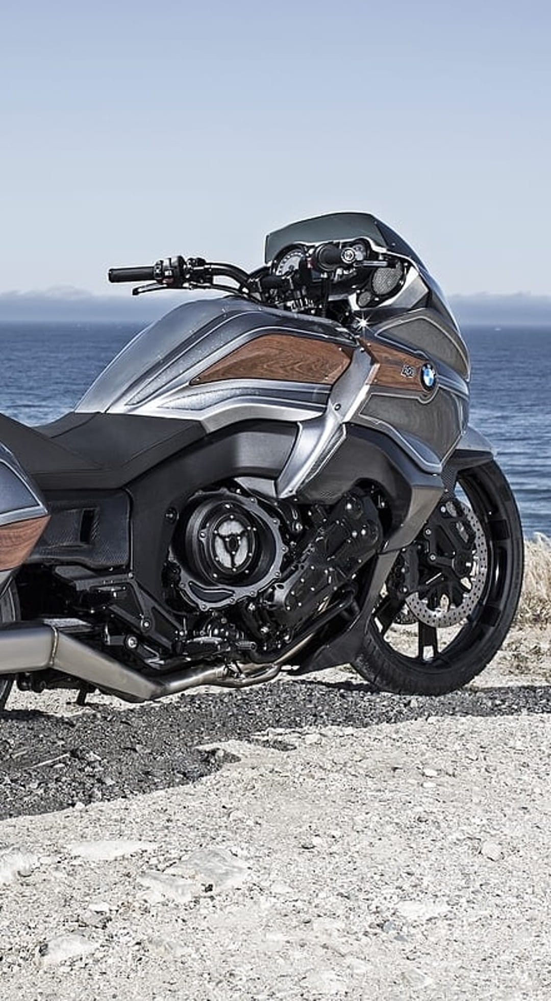 BMW Concept 101 motorcycle with wood accents parked on a sandy shoreline under a bright blue coastal sky.