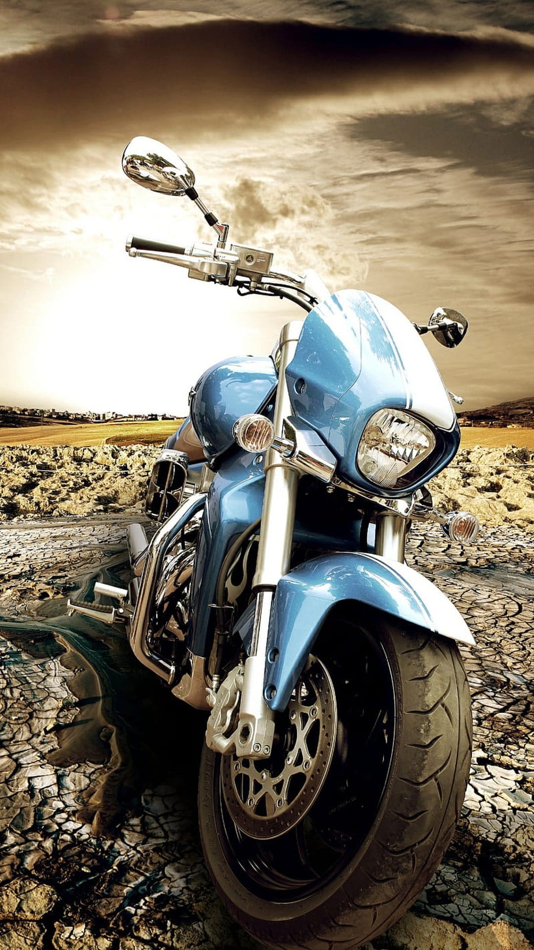 Blue cruiser motorcycle on cracked dry earth under a dark cloudy sky with golden sunset light at a low angle.