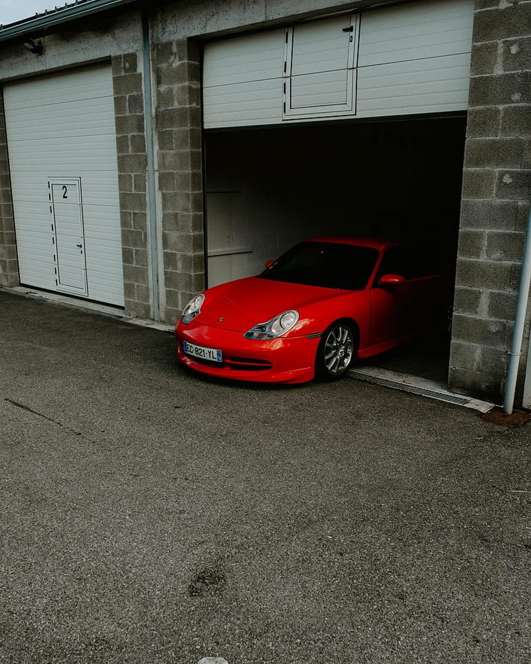 Red Porsche 911 Carrera parked in an open garage with grey brick walls and a dark textured asphalt driveway.