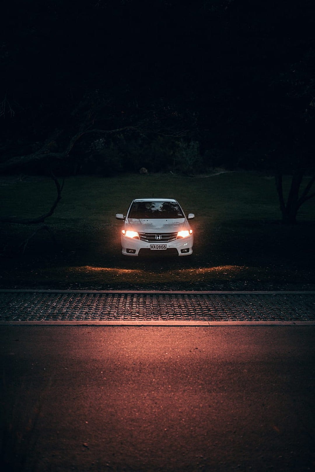 White Honda sedan with glowing headlights parked on grass at night, reflecting warm light on paved ground.
