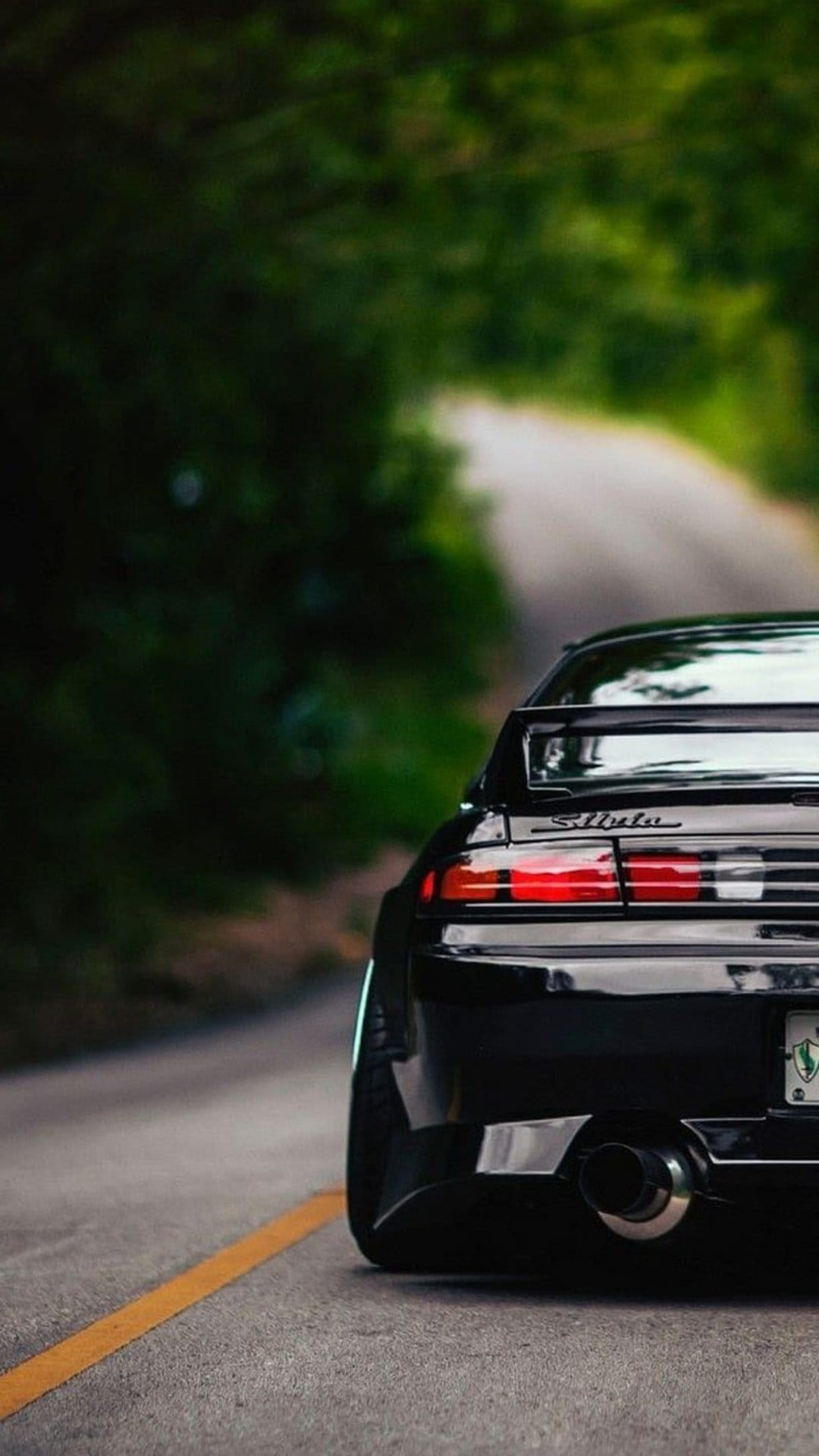 Black Nissan Silvia S14 rear view on a winding forest road with red taillights and a yellow lane line.