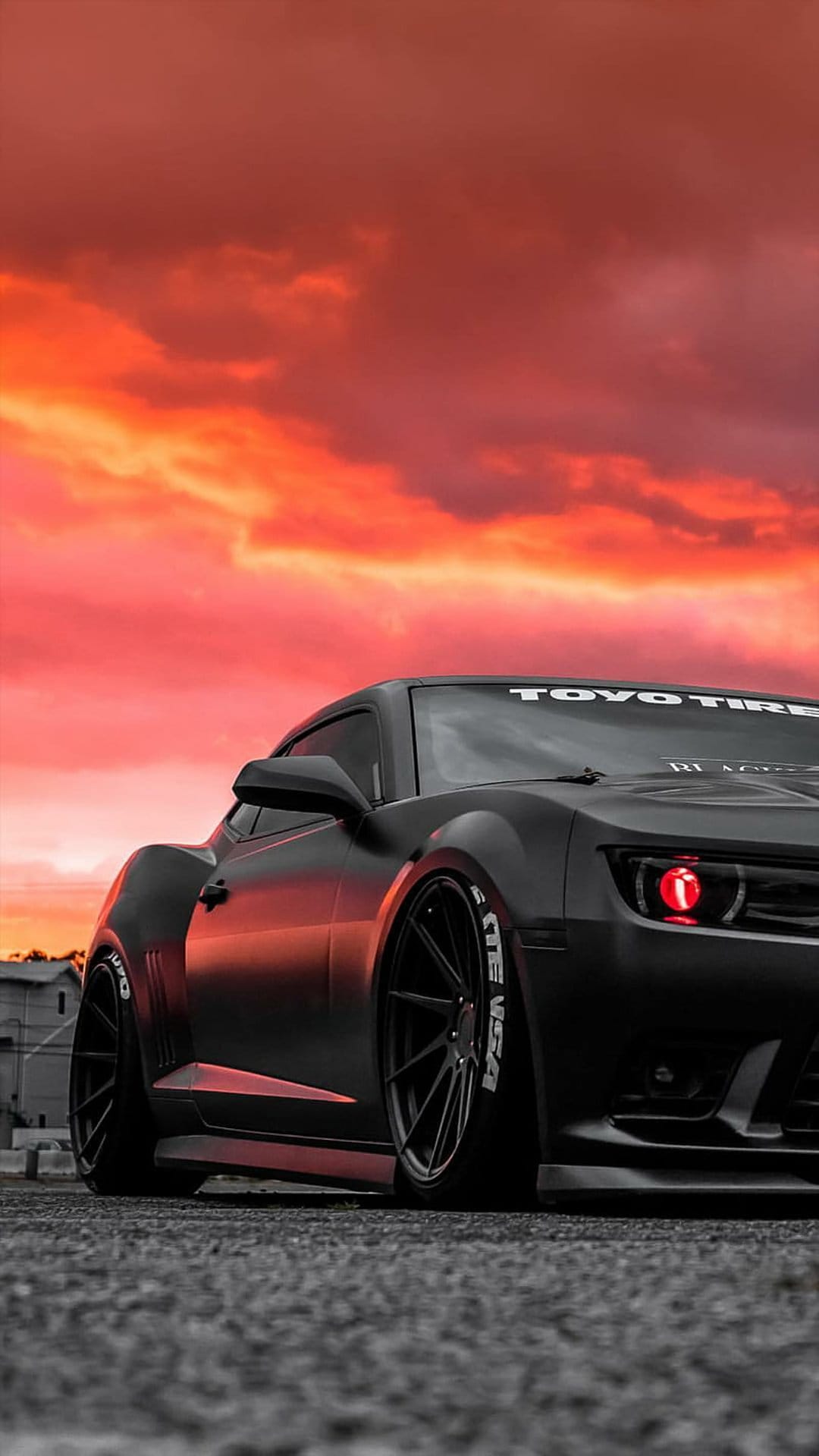 Black widebody Camaro with glowing red halo headlights parked on asphalt during a fiery orange sunset.
