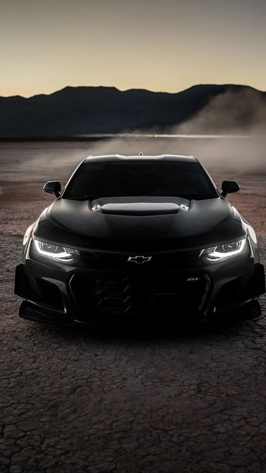 Black Chevrolet Camaro ZL1 kicking up dust on a dry desert road with mountains under a twilight sky.