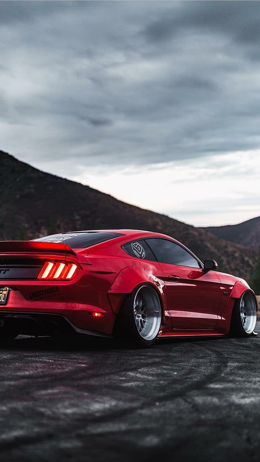 Red Ford Mustang with a widebody kit and silver rims on an asphalt road under moody, overcast mountain skies.