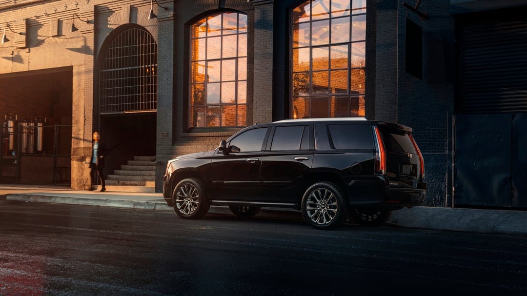 Black luxury SUV parked on a city street in front of a dark building with golden sunset reflections in the windows.
