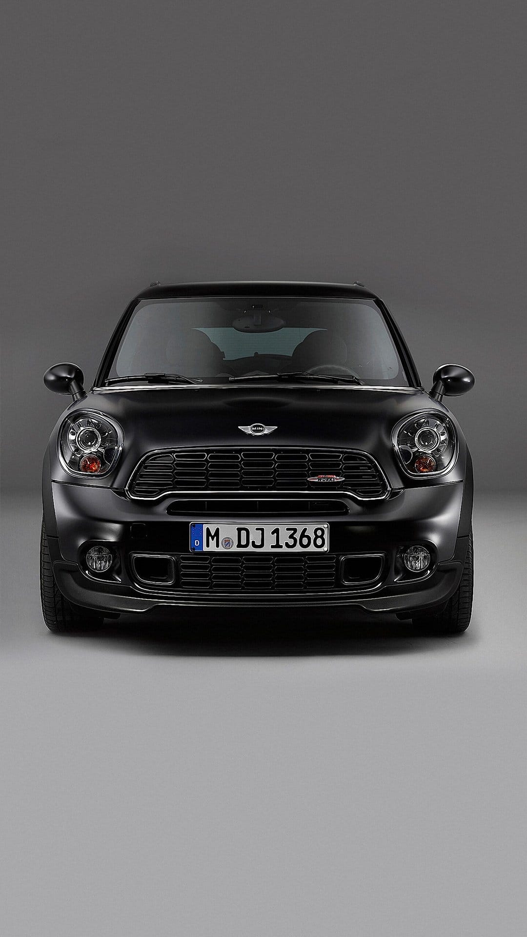 Matte black Mini John Cooper Works front view with headlights and red grille badge on a gray studio background.