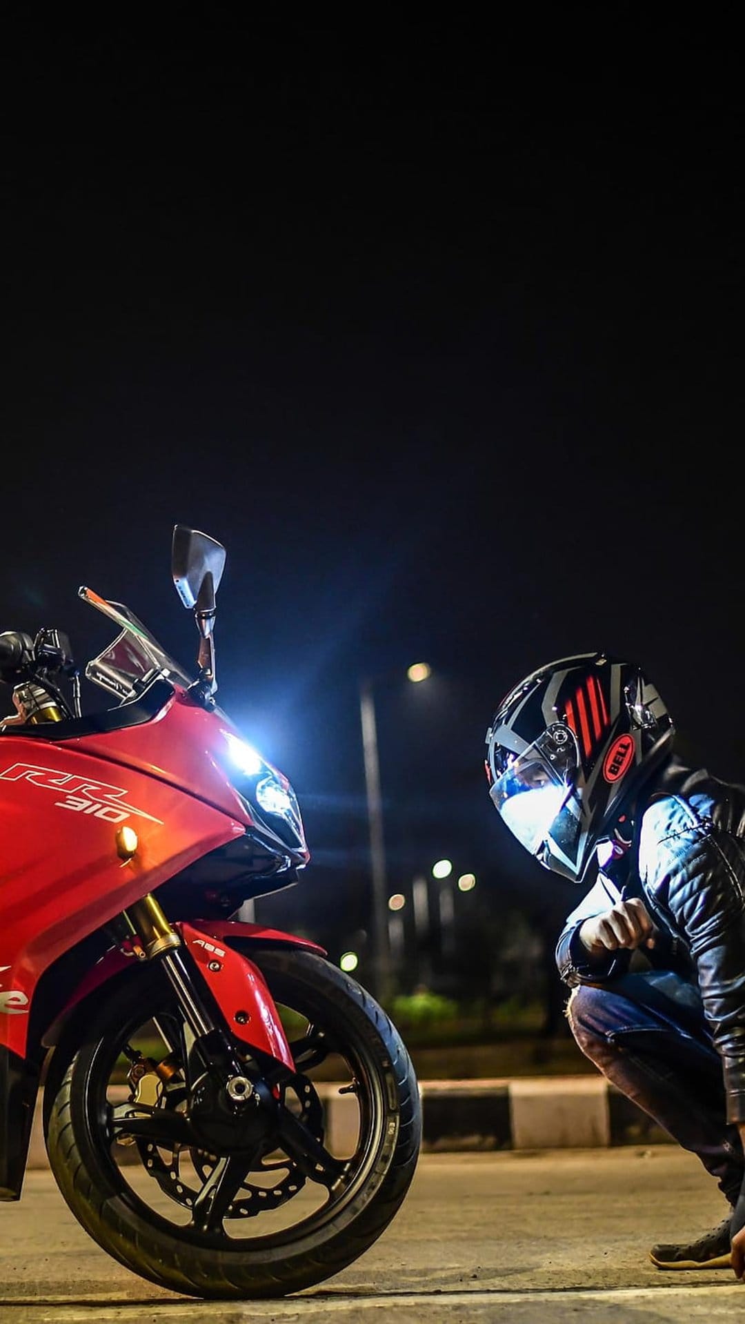 Motorcyclist in a leather jacket and helmet kneels next to a red sportbike with glowing headlights on a dark road.