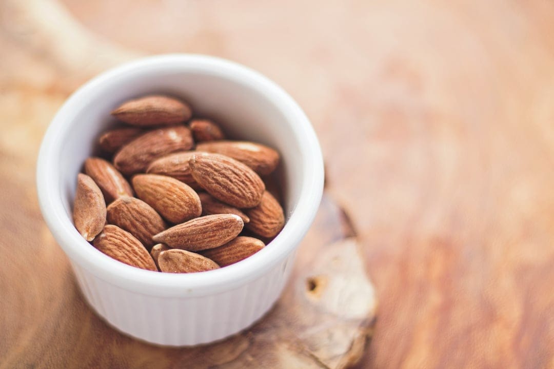 Whole almonds with textured brown skins sit in a white ceramic bowl on a blurred wooden table background.