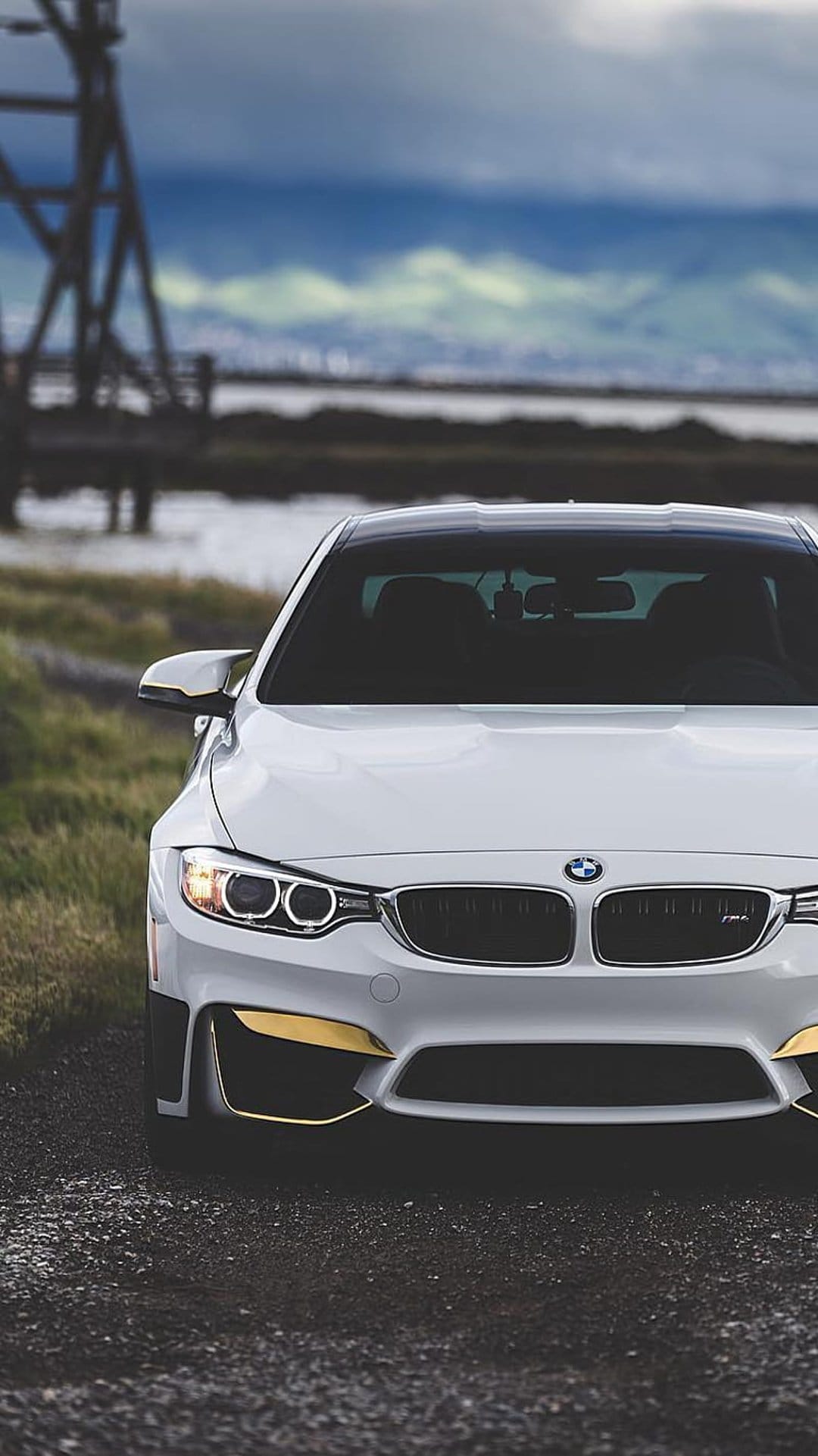 White BMW M4 coupe parked on a gravel road by a lake under a dark cloudy sky with golden bumper trim accents.