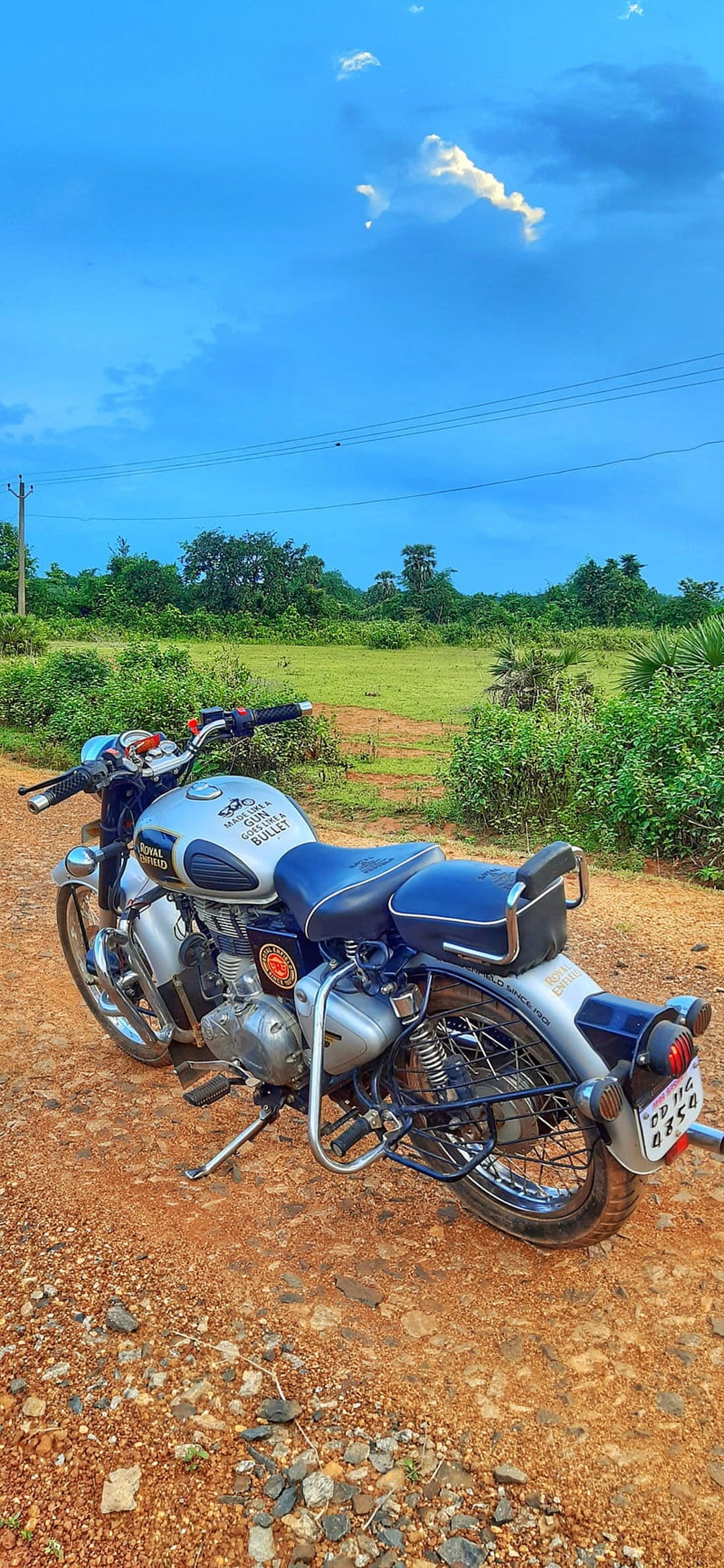 Royal Enfield Bullet motorcycle with a blue seat parked on a dusty rural road under a clear blue sky.