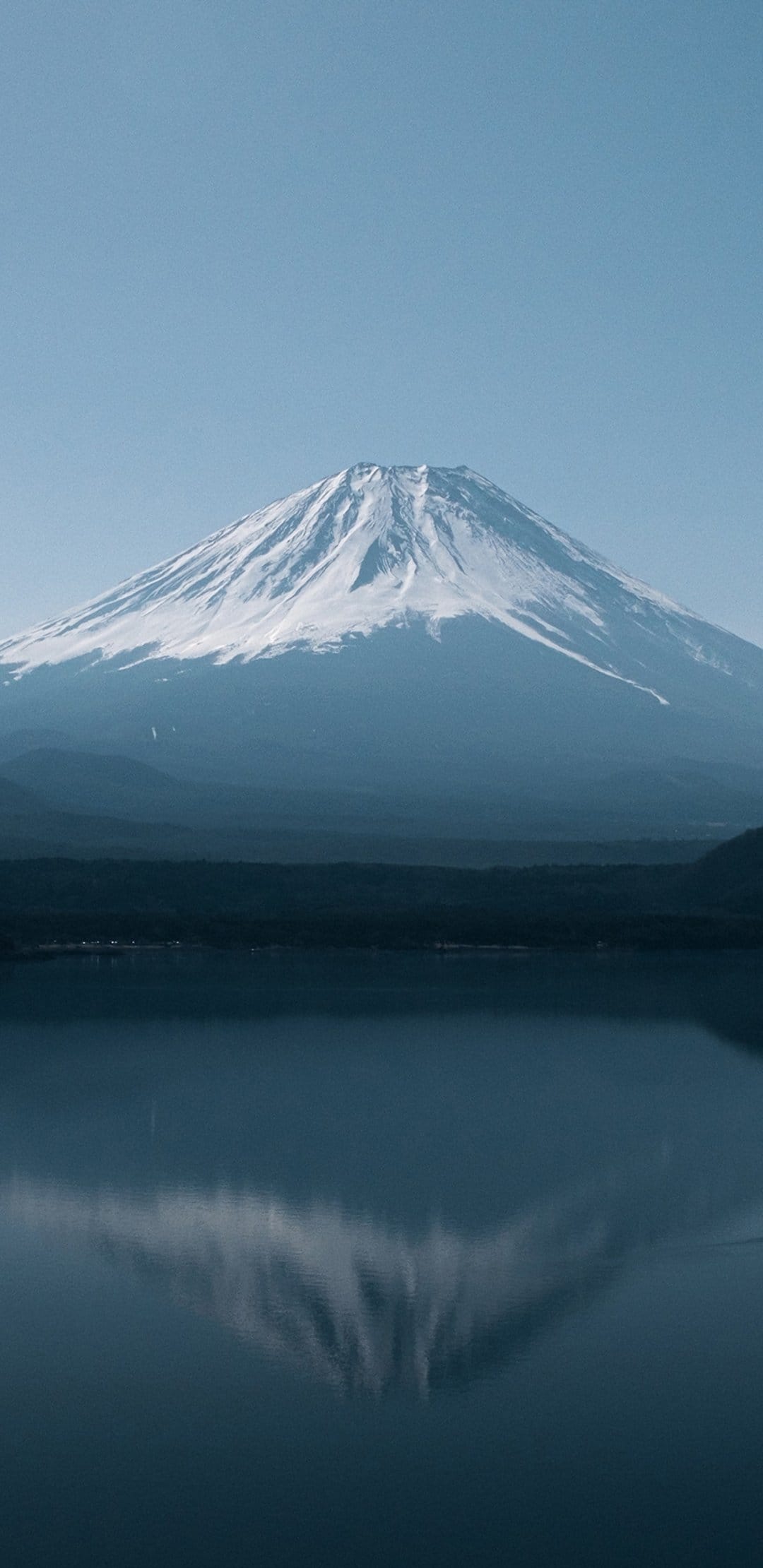 Mount Fuji with a snow-capped peak mirrors in the glassy blue water of Lake Kawaguchi under a clear morning sky.