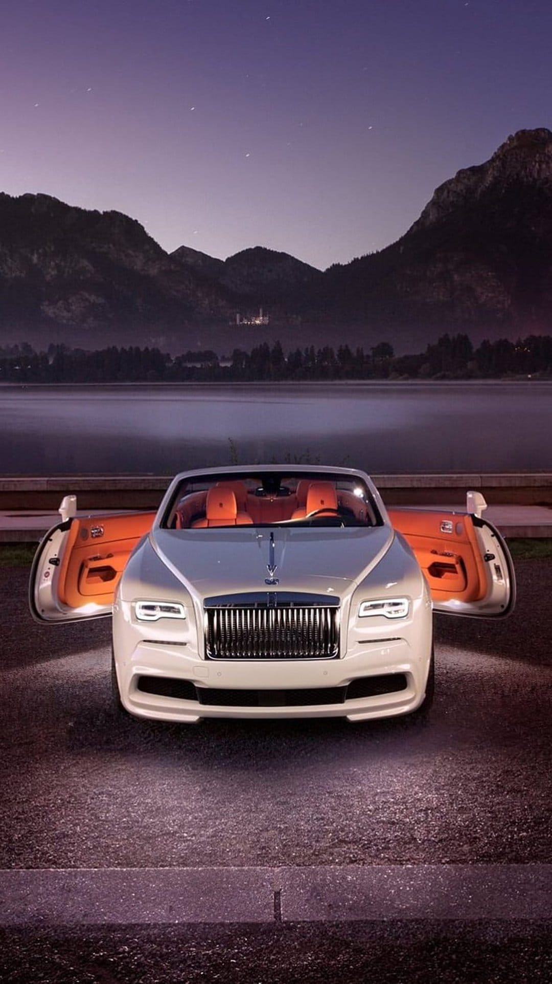 White Rolls-Royce convertible with orange seats parked near a mountain lake under a starry twilight sky.