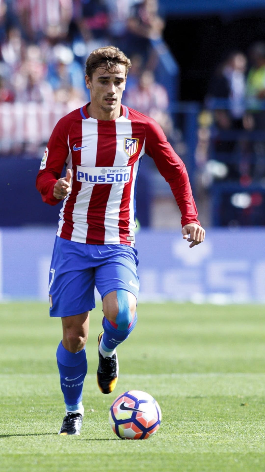 Antoine Griezmann in a striped Atletico Madrid kit dribbles a soccer ball on a green pitch with blurred fans.