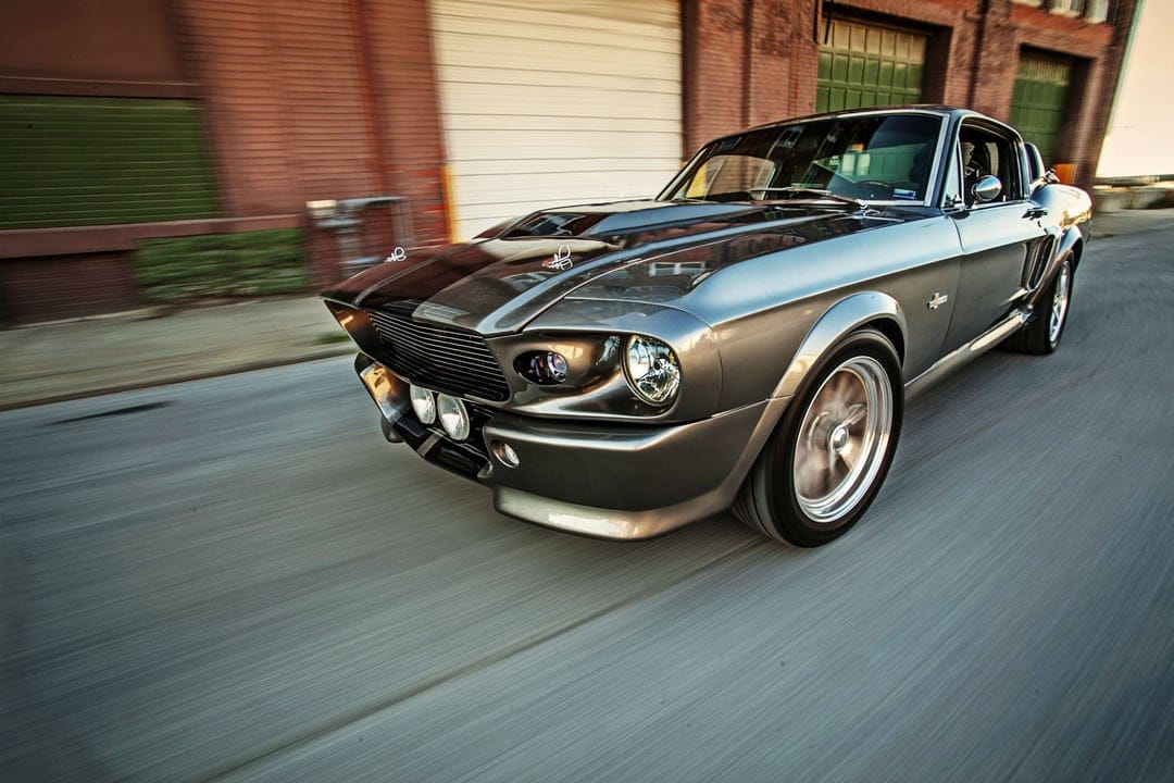Grey Mustang Shelby GT500 Eleanor driving fast past brick buildings with motion-blurred wheels and reflections.