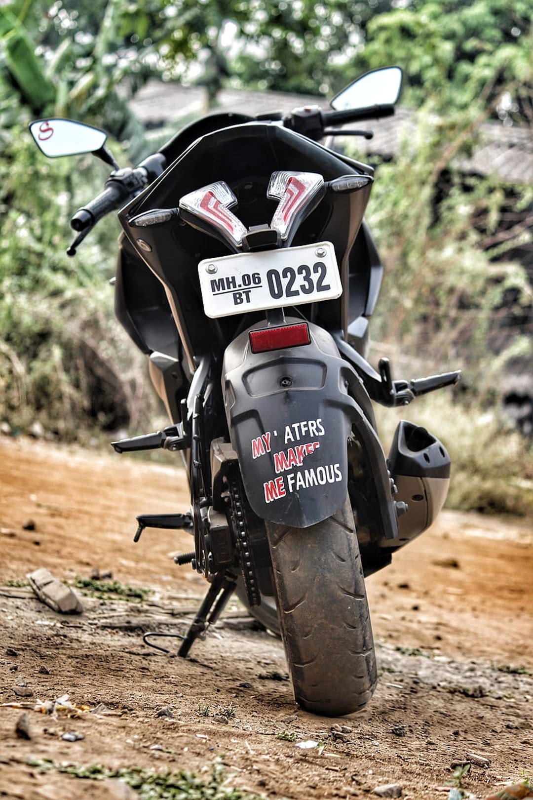 Rear fender of a black motorcycle showing a white license plate and red sticker on a blurred forest road.