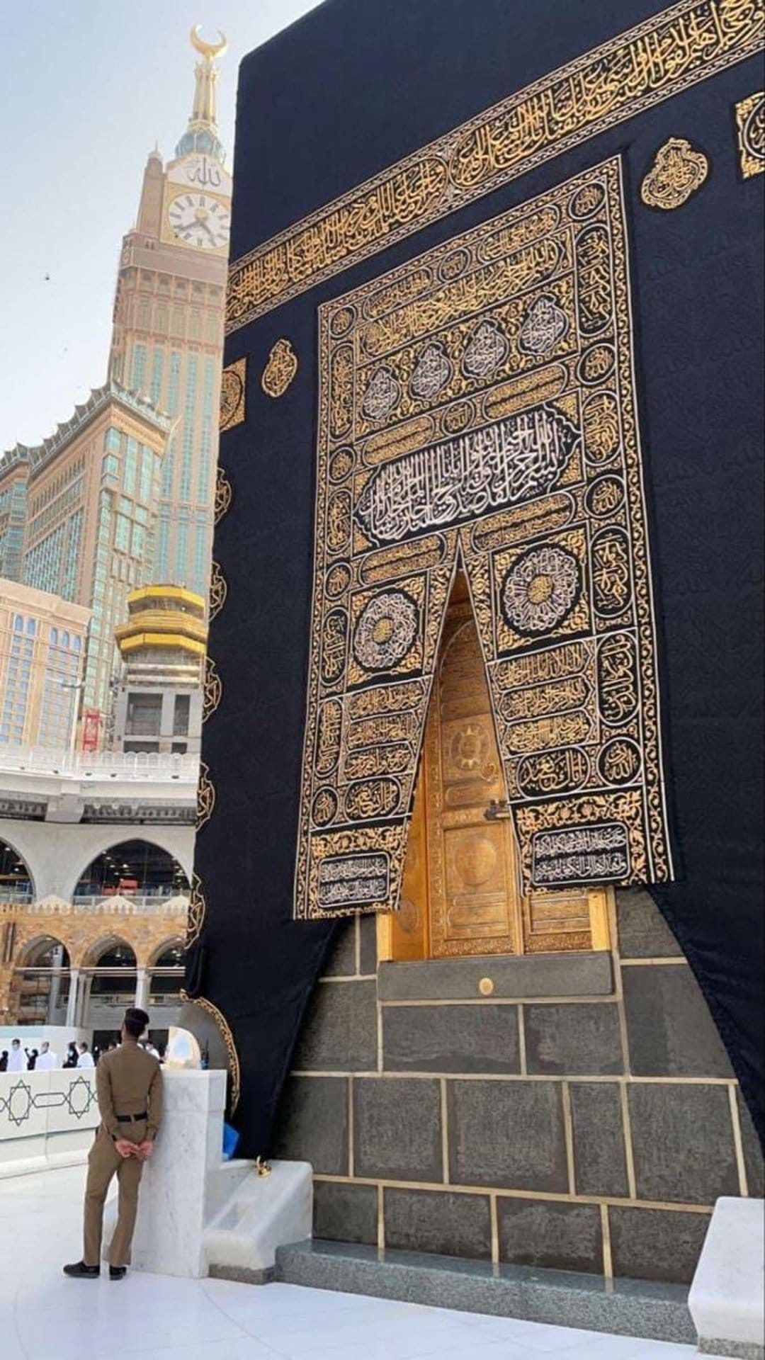 Kaaba door with golden Arabic calligraphy stands before the distant Makkah Clock Tower under a clear sky.