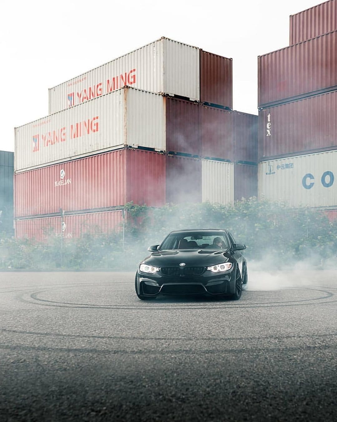 Black BMW M4 drifting amid thick white tire smoke in front of stacked industrial shipping containers.