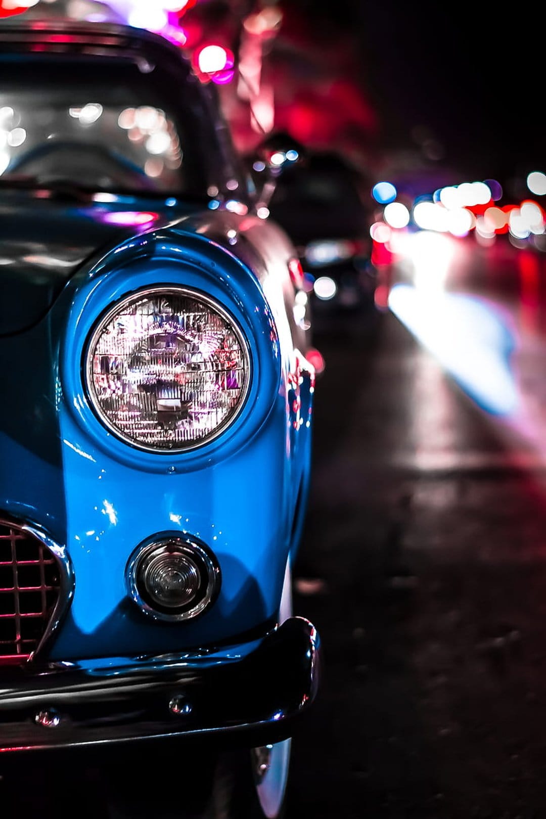 Round glass headlight of a vintage blue car reflecting city lights against a background of colorful bokeh.
