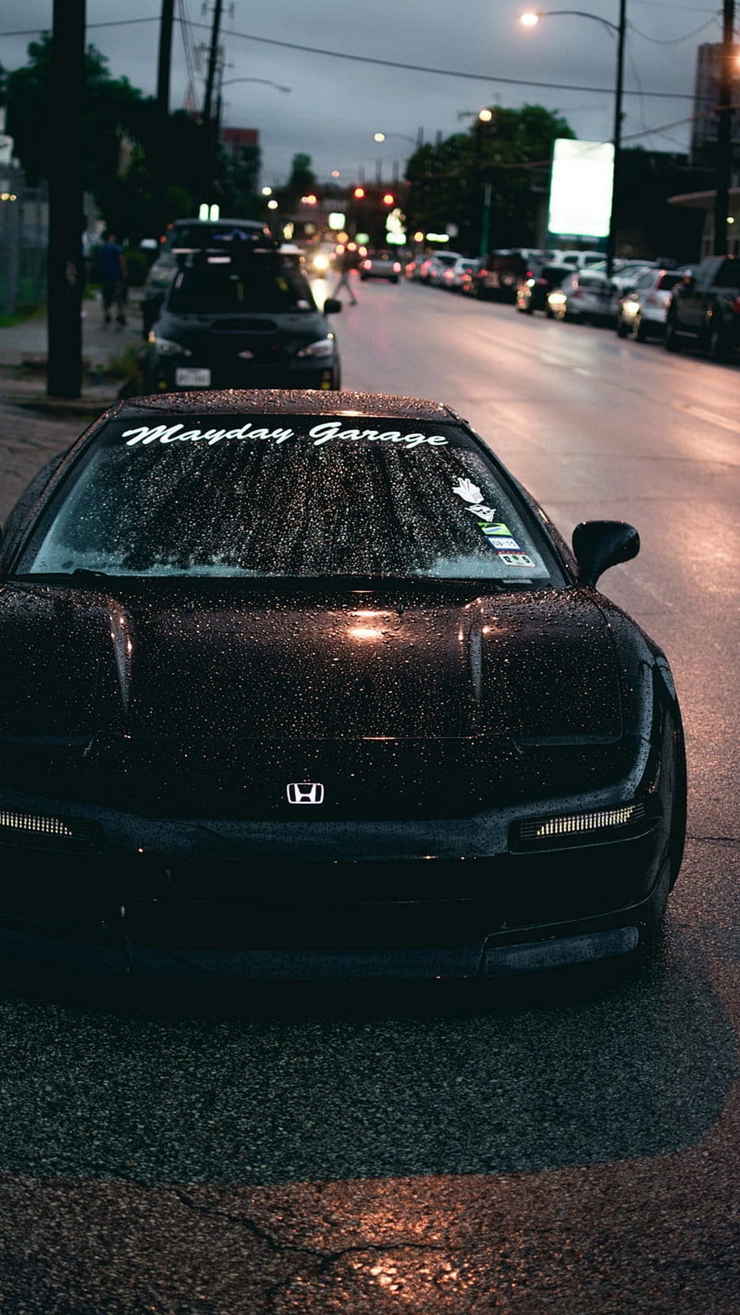 Honda NSX sports car with a windshield banner parked on a wet asphalt road under urban night lights.