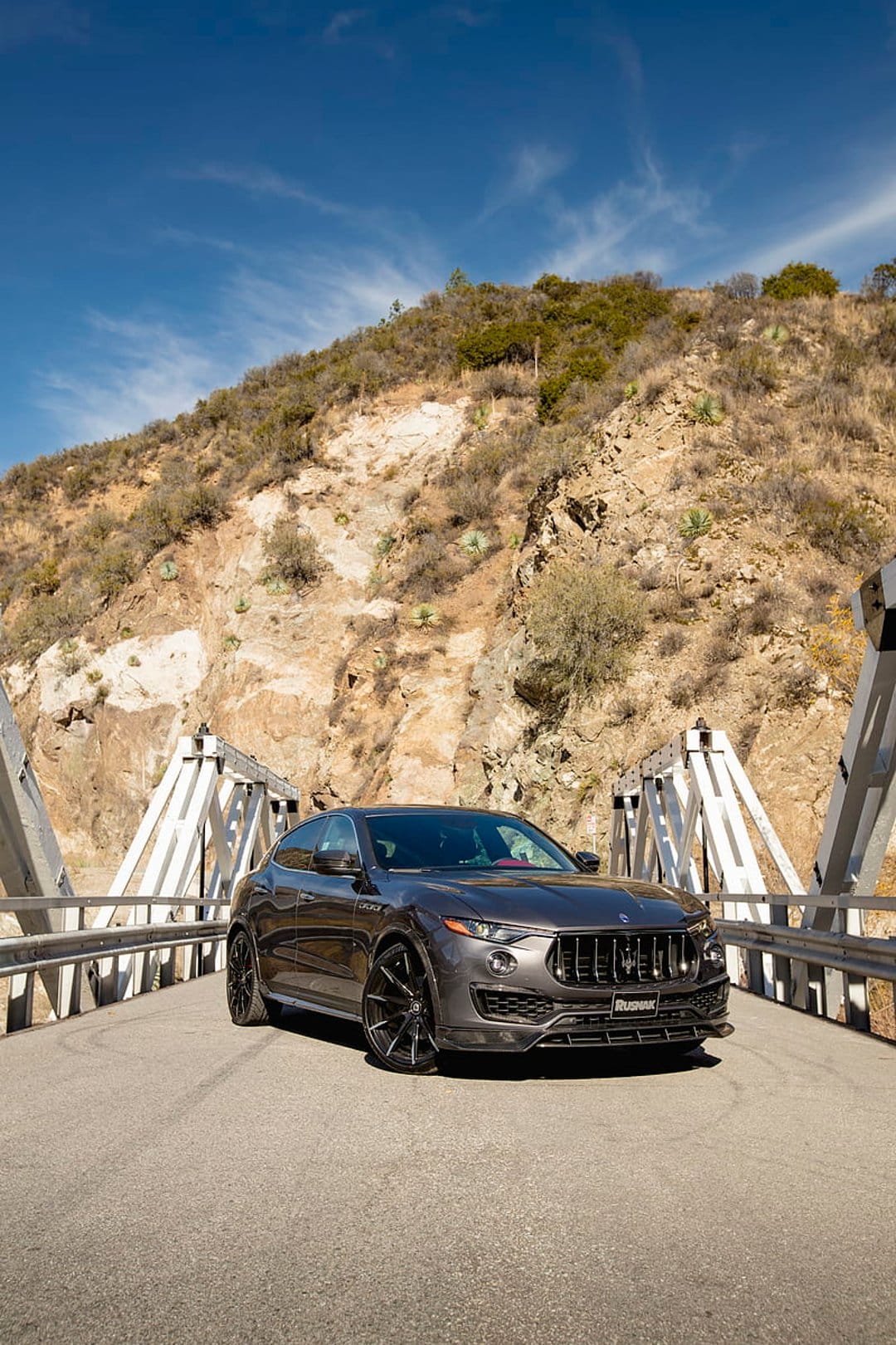 Maserati Levante SUV parked on a white metal bridge with a dry mountain range under a clear blue sky.
