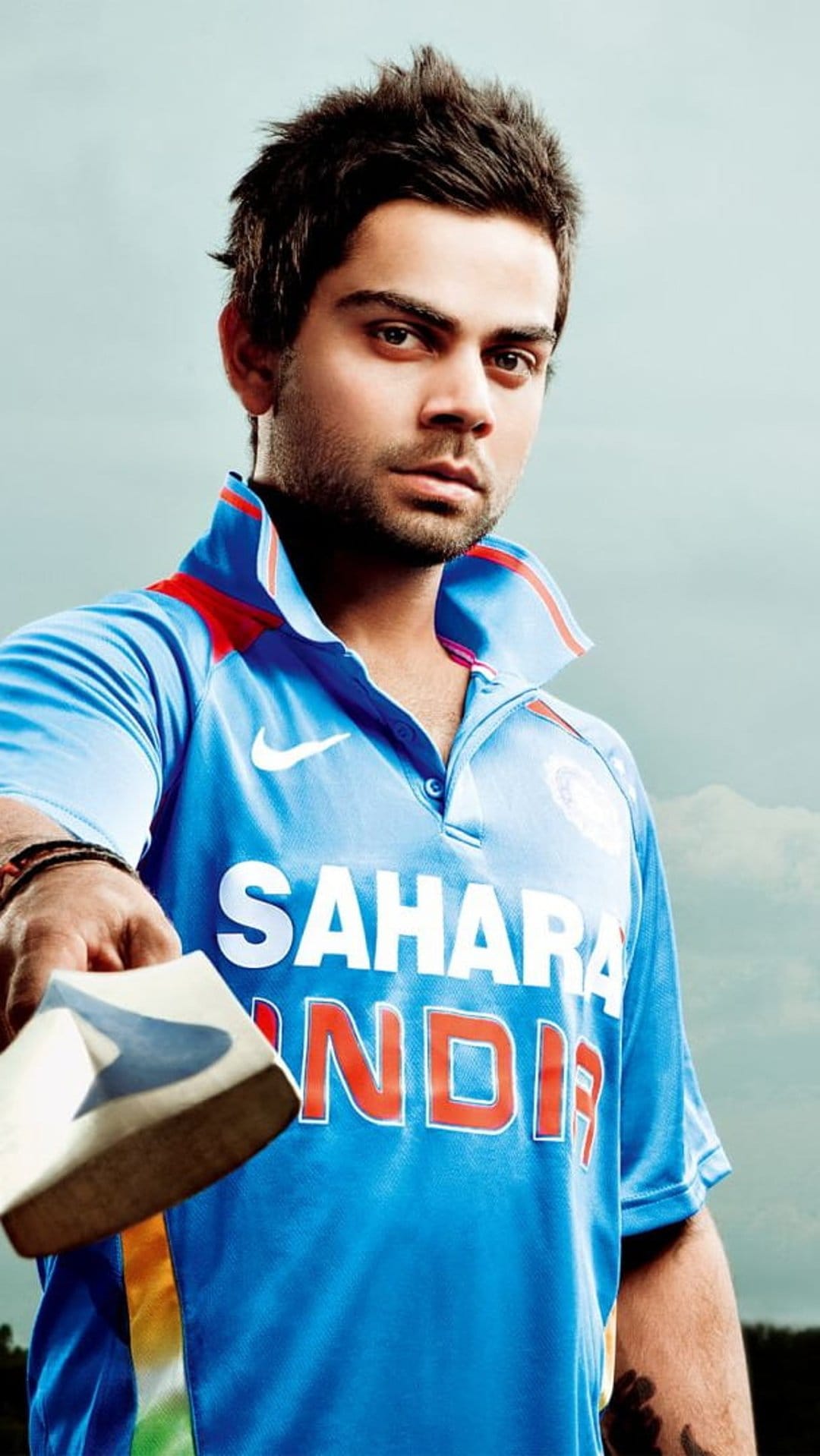 Virat Kohli in a blue Indian cricket jersey looking forward with an intense expression against a blurred background.