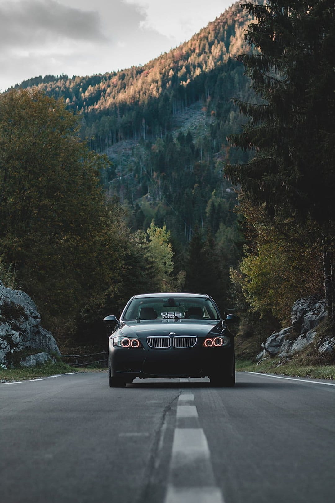 Black BMW sedan drives on a winding mountain road surrounded by golden autumn forests under a cloudy sunset sky.
