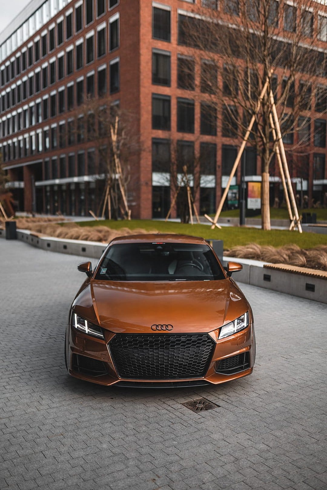 Orange Audi TT coupe with an aggressive front grille parked on a cobblestone street in front of a brick building.