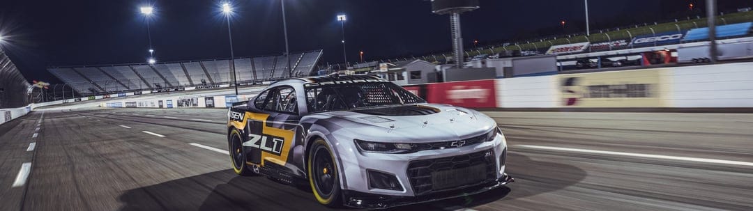 Chevrolet Camaro ZL1 race car speeding on a track at night under bright stadium floodlights with motion blur.