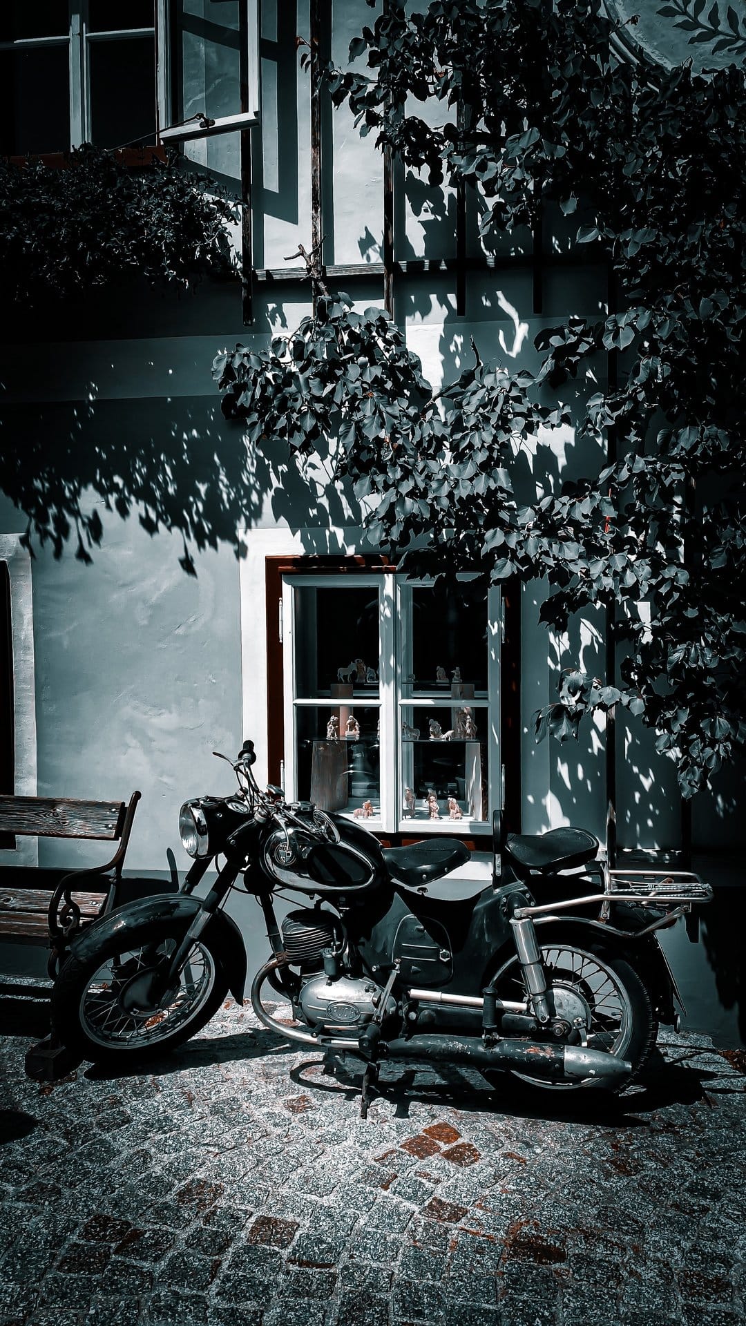 Vintage black motorcycle on a cobblestone path beside an ivy-covered building with dappled sunlight shadows.