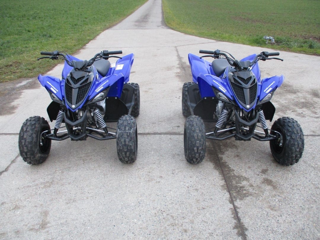 Two blue Yamaha Raptor ATVs parked side-by-side on a narrow concrete path between lush green grass fields.