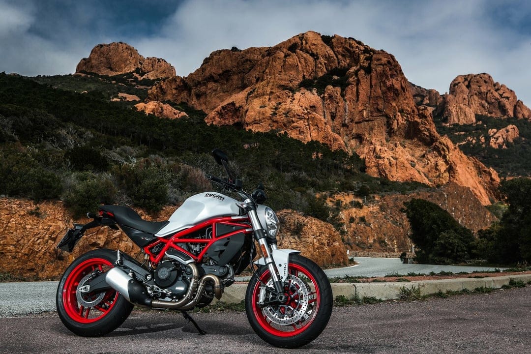 White Ducati Monster motorcycle with red frame parked on a road with red rocky mountains in the background.