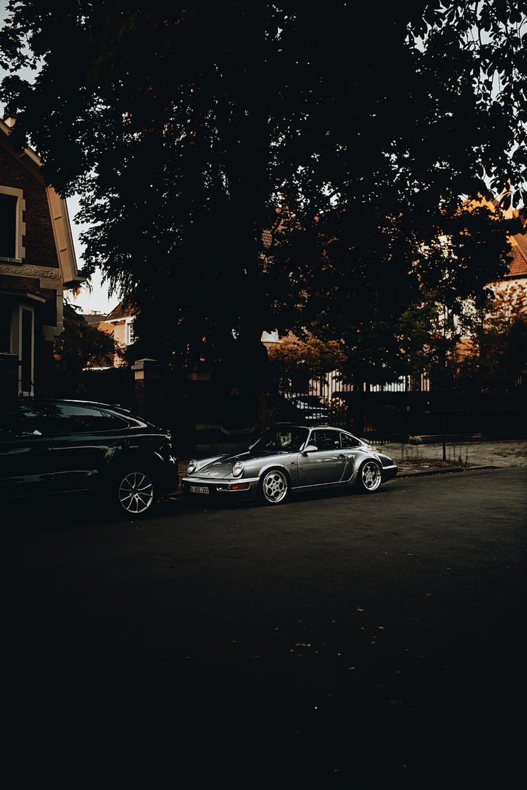 Silver vintage Porsche 911 parked on a street at dusk under a tree beside a dark SUV during golden hour.