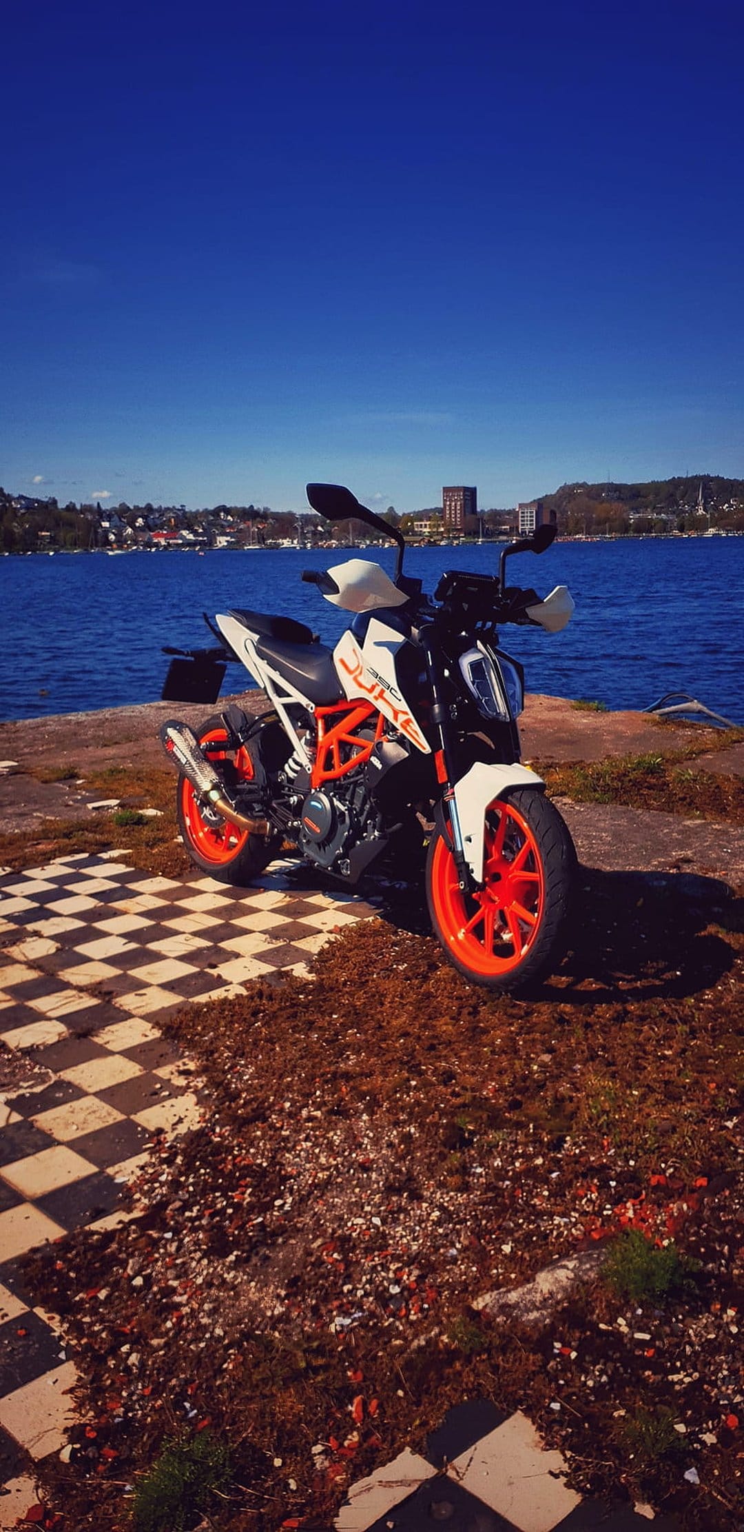 KTM Duke motorcycle in orange and white parked on a rocky shore beside a bright blue lake and distant hills.