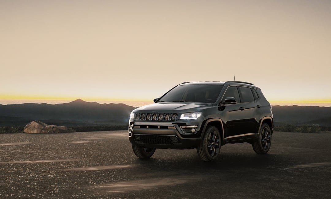 Black Jeep Compass SUV parked on a dark surface with glowing headlights against a sunset mountain range.