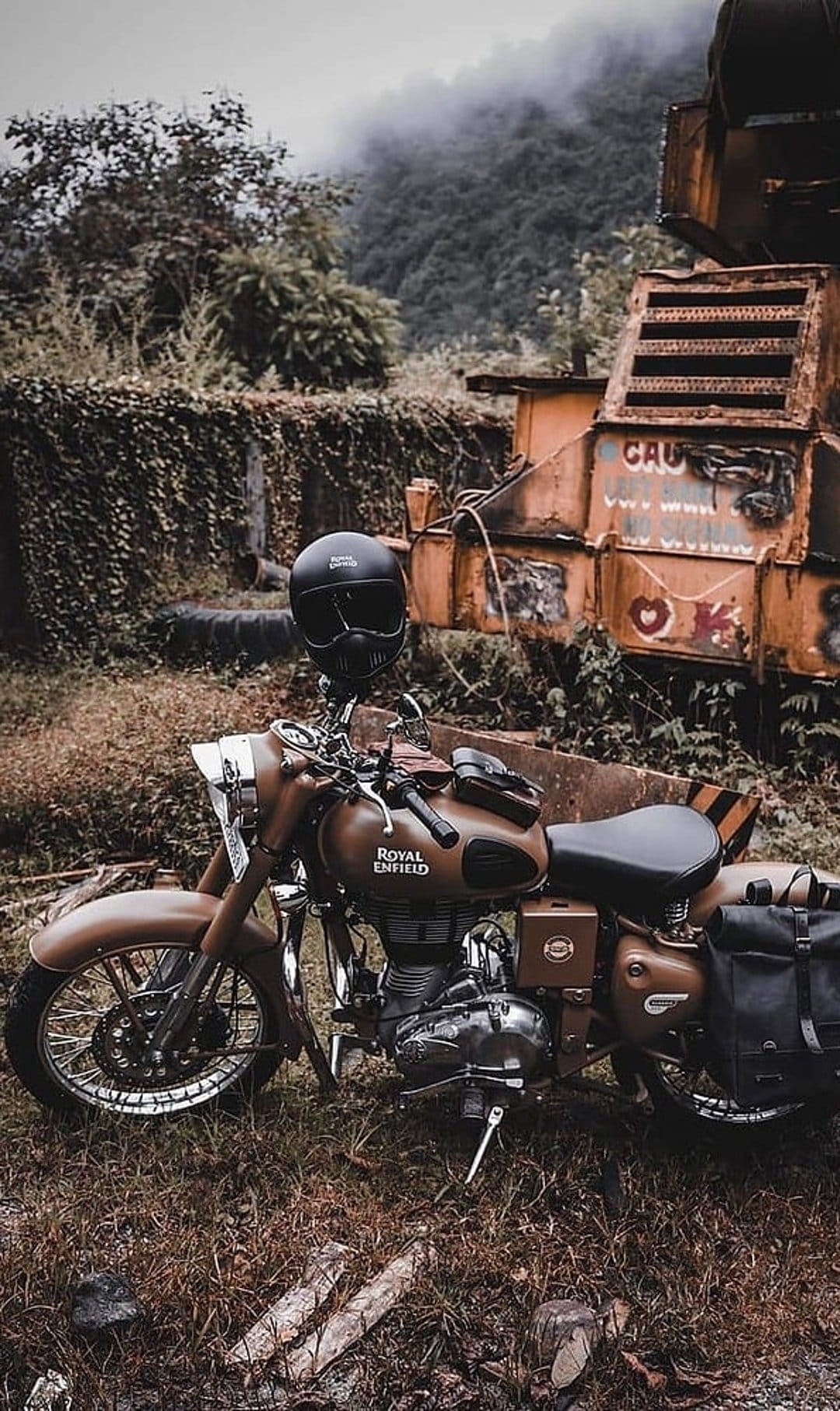 Brown Royal Enfield motorcycle with a black helmet parked near a rustic building and foggy forest hills.