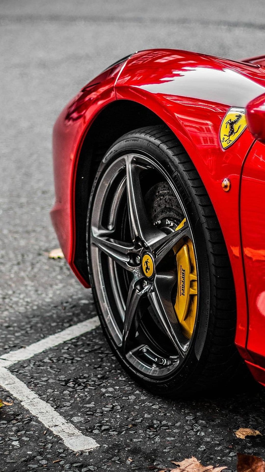Red Ferrari fender and alloy wheel with a yellow brake caliper on asphalt with fallen leaves in a macro view.