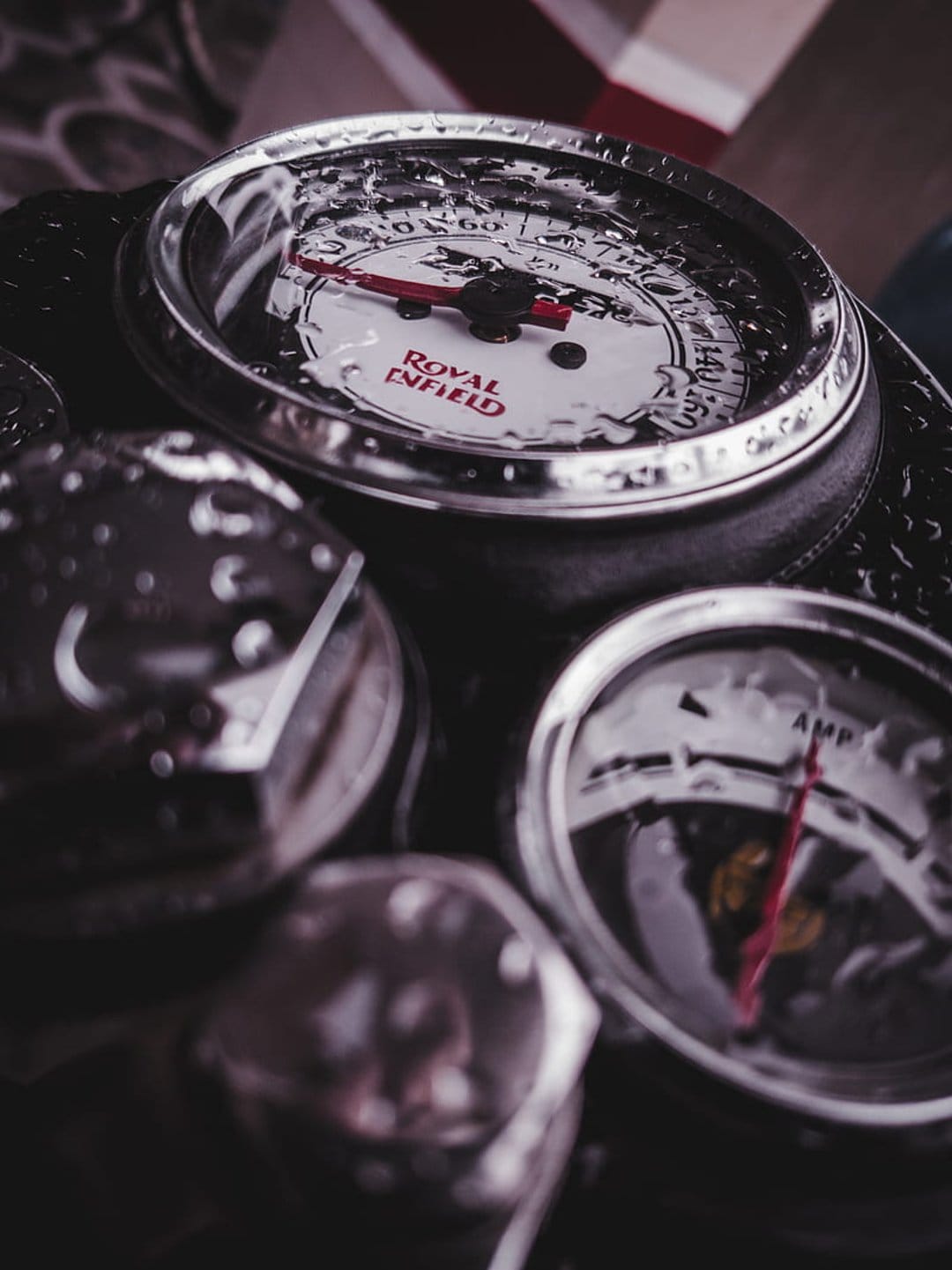 Royal Enfield speedometer and ammeter covered in small water droplets with a weathered chrome dashboard finish.
